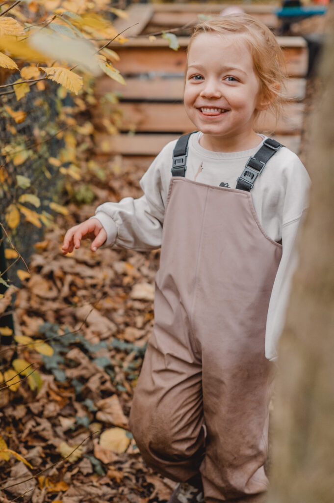 Ein Kindergartenkind trägt eine Matschhose und steht in einem Blätterberg. Es ist Herbst und das Mädchen lacht in die Kamera.
