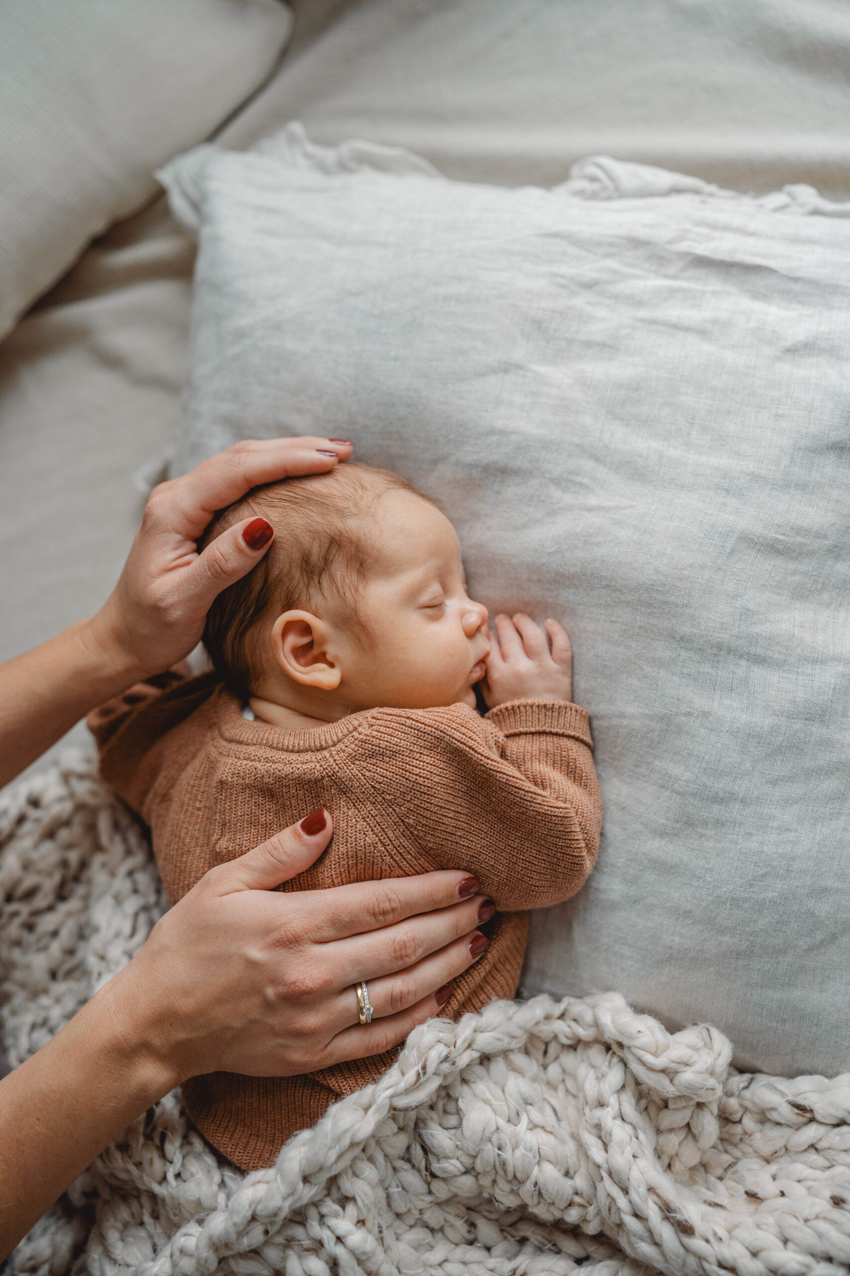 Ein neugeborenes Baby liegt schlafend auf einem Kissen. Dazu sieht man eine Hand, die den Kopf vom Baby liebevoll hält und eine Hand liegt auf dem Rücken.